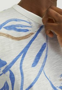 Vit t-shirt med kort ärm som har blå och bruna abstrakta penseldragmönster. Rundhalsdesign med ett mjukt, texturerat tyg.