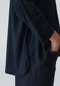 Maglia a maniche lunghe blu navy con vestibilità rilassata e orlo arrotondato, realizzata in tessuto morbido. Mano visibile in tasca, abbinata a jeans di denim scuro.