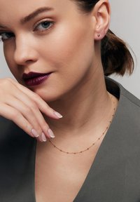 Złota naszyjnik z równomiernie rozłożonymi małymi kulkami, noszony na gładkiej karnacji. Modelka ma ciemne usta i neutralny, szary strój z dekoltem w kształcie litery V.