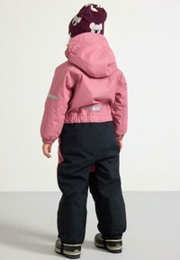 Giacca impermeabile rosa con cappuccio e dettagli riflettenti, abbinata a pantaloni impermeabili neri. Stivali scuri completano l'outfit.