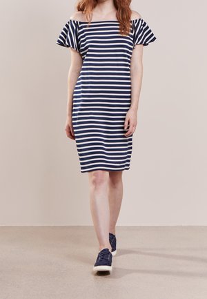 Woman walking wearing a navy and white striped off-shoulder dress with navy sneakers on a plain beige background.