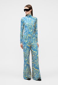 Woman wearing blue and yellow abstract patterned long-sleeve top and wide-leg pants, black oversized sunglasses, standing against white background.