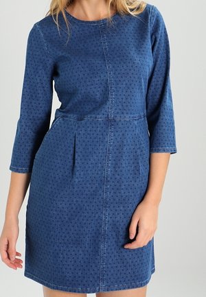 Frau trägt ein blaues, knielanges Kleid mit 3/4-Ärmeln und dezentem Polka-Punktmuster, steht vor einem einfarbigen hellen Hintergrund.