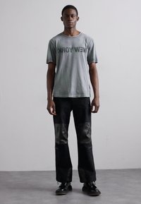 Graues Baumwoll-T-Shirt mit "NEW YORK" in Dunkelgrün bedruckt, kombiniert mit schwarzen Hosen, die glänzende Leder-Patches und Mesh-Akzente aufweisen.
