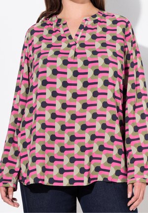 Bluzka z długim rękawem w geometryczny wzór w kolorach różowym, oliwkowym, czarnym i beżowym, z dekoltem w kształcie litery V, noszona z ciemnymi spodniami.