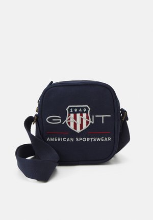 Sac bandoulière carré bleu marine avec sangle réglable, logo GANT, emblème en forme de bouclier avec "1949" et texte "American Sportswear" sur le devant.