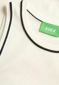 Hvid tanktop med en sort kant rundt om halsudskæringen, der har et grønt mærke med teksten "JJXX". Blødt stof.