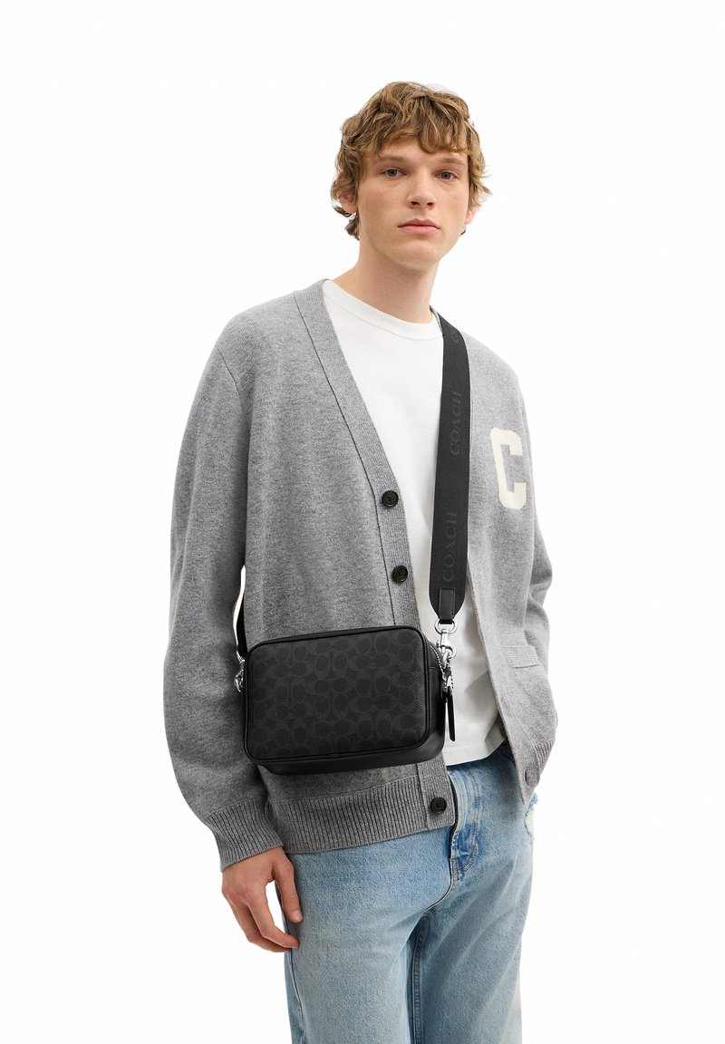 Joven con cárdigan gris y camisa blanca, con bolso bandolera negro estampado y vaqueros azul claro sobre fondo blanco.