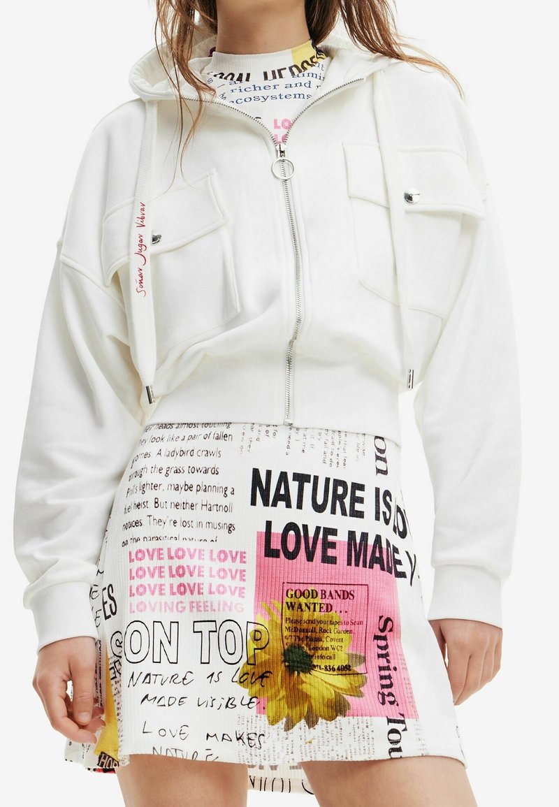 Hoodie bianco corto con due tasche frontali e zip. Abbinato a una gonna con motivo misto di testo e fiori nei colori rosa, giallo e nero.
