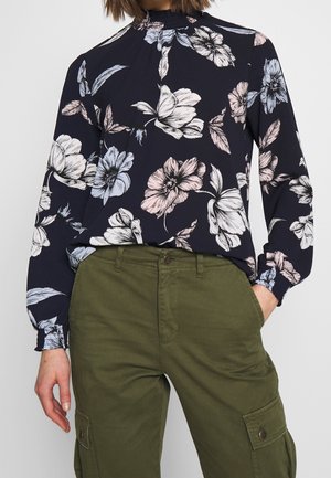 Vrouw draagt een marineblouse met grote roze en witte bloemenprints en olijfgroene high-waisted cargobroek met zakken.
