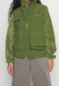 Zelená vesta vyrobená z měkkého fleecu, s kapsami na zip a klopou, oranžovými akcenty na zipu a síťovinou v oblasti ramen.