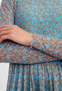 Modrá šaty se skvrnitým vzorem v hnědé a tyrkysové barvě. Mají dlouhé rukávy se zkroucenými okraji a designem plisované sukně. Hladká textura.