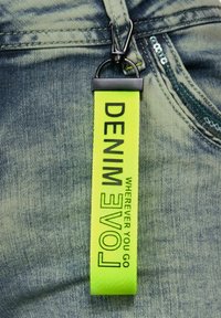 Etichetta in tessuto giallo brillante con la scritta "DENIM" e "LOVE", dotata di un gancio nero, posizionata su un materiale in denim blu.