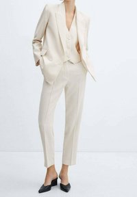 Béžový oblek na míru s jednoradovým sakem, odpovídající vestou a slim-fit kalhotami. Má hladkou látku a ostré klopy. Černé muly.
