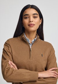 Maglione marrone con chiusura a zip e maniche lunghe, caratterizzato da una texture a costine, un piccolo logo sul petto e un colletto sopra una camicia azzurra chiara.