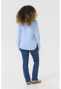 Helles blaues Hemd mit langen Ärmeln und abgerundetem Saum, kombiniert mit dunkelblauben Jeans und hellbraunen Schuhen. Texturiert, weicher Stoff, lässige Passform.