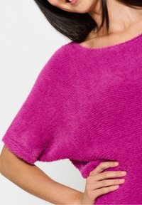 Luddig, oversized topp i klar rosa. Har en vid halsringning och korta ärmar, vilket visar en mjuk textur med ett subtilt mönster.