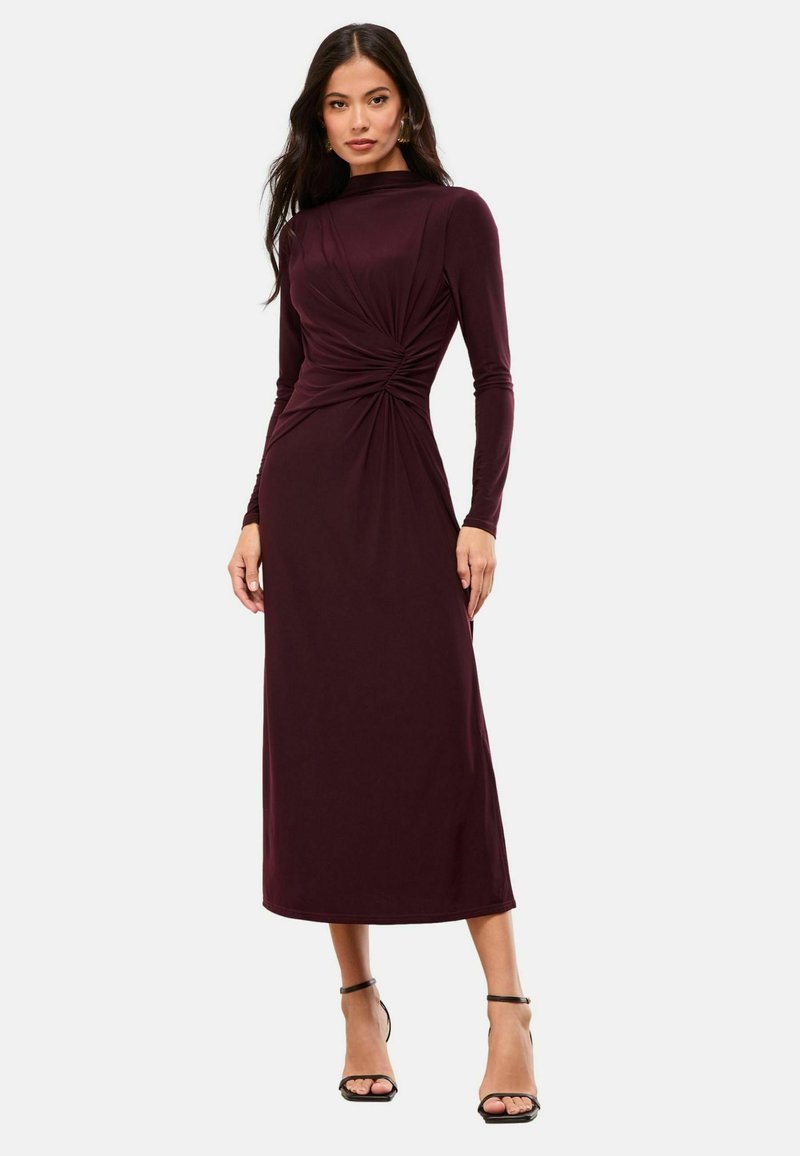 Rochie long-sleeve burgundy cu un design răsucit la talie, realizată dintr-un material neted, până la glezne, asortată cu sandale negre cu curele.