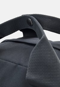 Gros plan sur une poignée de sac en tissu noir sécurisée par un bouton-pression noir et renforcée par des sangles tissées texturées.