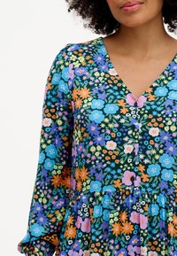Robe fleurie avec un décolleté en V, des manches longues et une jupe à volants. Le tissu présente un motif coloré de fleurs bleues, oranges et violettes sur fond vert.