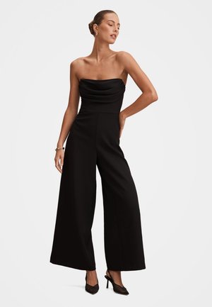 Femme portant une combinaison noire bustier à jambes larges et des talons noirs, posant avec une main sur la hanche sur un fond blanc.