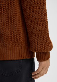 Maglione lavorato a maglia con texture in un caldo marrone, con polsini e orlo a coste. Indossato sopra un tessuto scuro, che mette in evidenza i dettagli delle mani e il motivo dei punti.