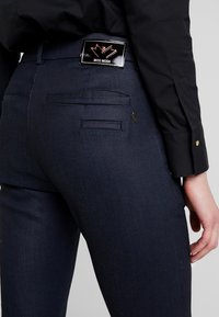 Pantalones de color azul oscuro con una textura suave, que cuentan con una etiqueta de metal negro de marca y dos bolsillos traseros. La manga de una camisa negra es parcialmente visible.