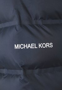 Gros plan sur un tissu sombre avec des coutures horizontales et "MICHAEL KORS" imprimé en lettres blanches.