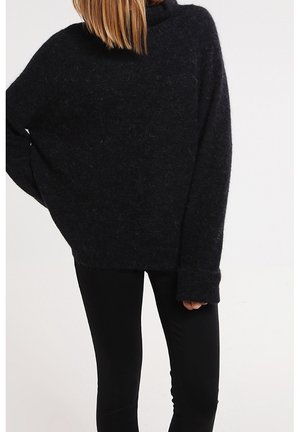 Person trägt einen lockeren, flauschigen schwarzen Pullover mit langen Ärmeln und eng anliegenden schwarzen Hosen vor einem schlichten hellen Hintergrund.