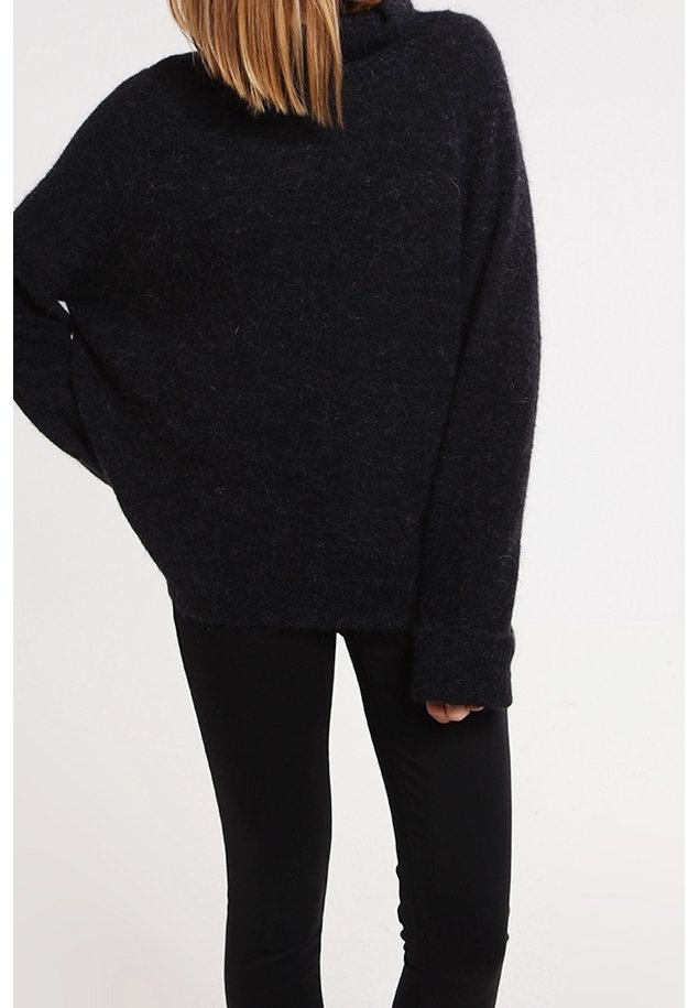 Personne portant un pull noir ample et duveteux à manches longues et un pantalon noir ajusté sur un fond clair uni.