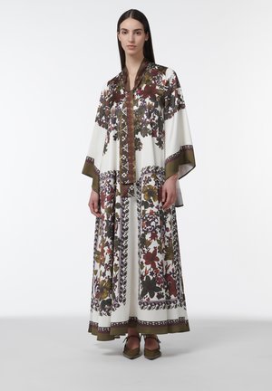 Mujer vestida con un largo vestido blanco de mangas anchas y estampados florales multicolores, combinado con zapatos puntiagudos en tono verde oliva.