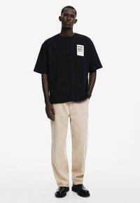 Black cotton t-shirt with short sleeves and a square graphic patch, paired with light beige wide-leg pants and black shoes.