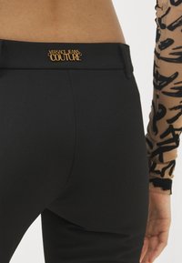 Juodos kelnės su tekstūruotu audiniu, ant juosmens puikuojasi auksinis "Versace Jeans Couture" logotipas. Detalus galinis vaizdas.