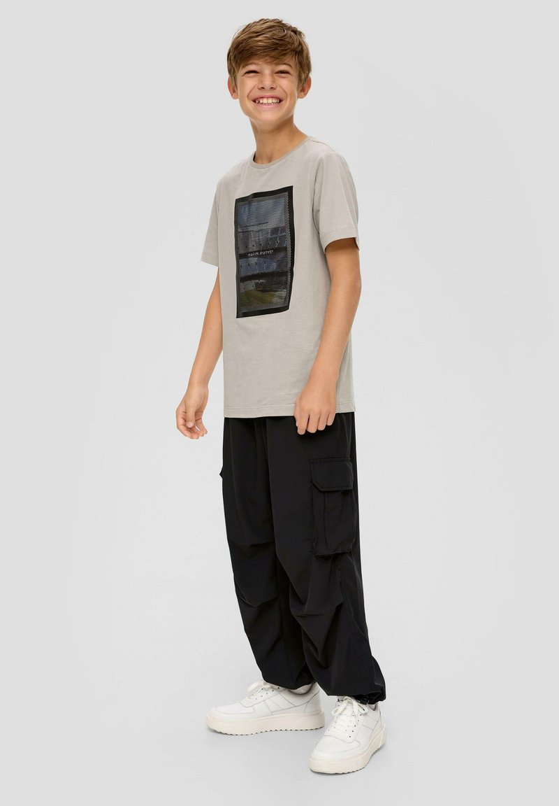 Hellgraues T-Shirt mit einem strukturierten schwarzen quadratischen Design, das eine Landschaft darstellt, kombiniert mit lockeren schwarzen Cargo-Hosen und weißen Sneakers.
