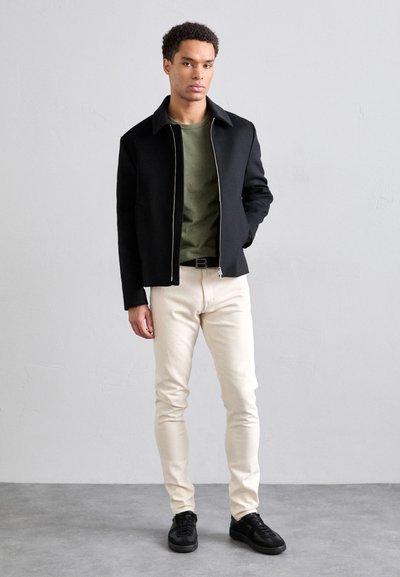 Černá bunda s límcem a zipem, zelená košile, krémové džíny. Černé boty, jednoduchý design, slim fit, minimální kovové prvky.