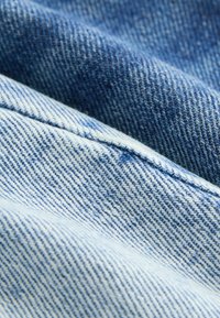 Denimstoff in unterschiedlichen Blautönen, mit einem subtilen Köpergewebe und kontrastierenden helleren Streifen. Glatte Textur mit sichtbaren Nähten.