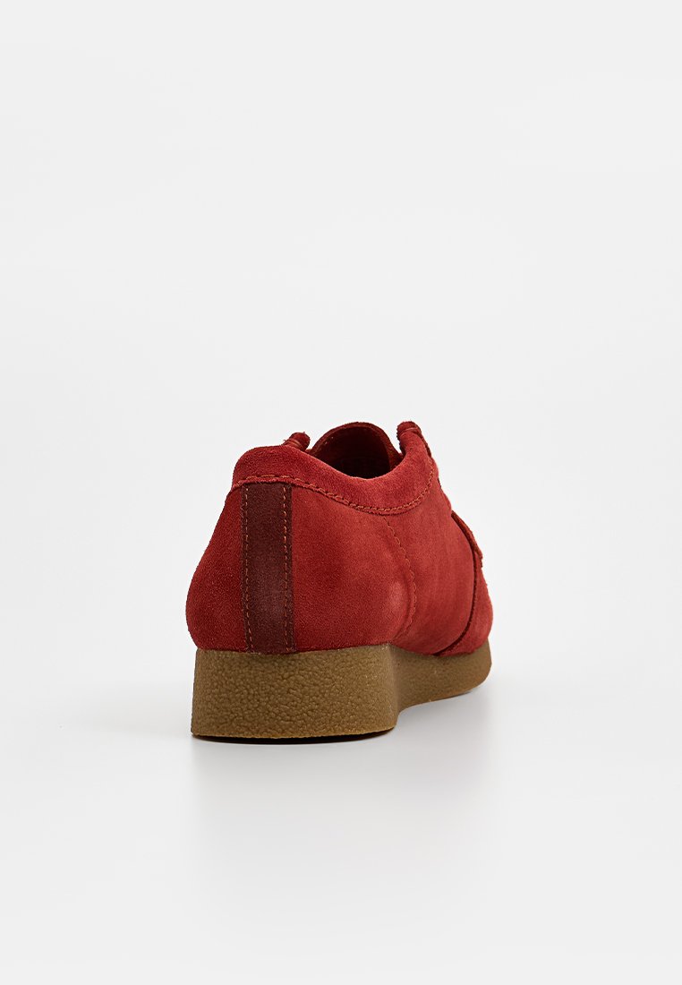 Roter Wildlederschuh mit glatter Textur, niedrigem Absatz und rundem Zehenbereich. Die Rückseite hat einen braunen Akzent und detaillierte Nähte entlang der Kanten.