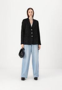 Blazer texturé noir avec des boutons en argent, associé à un jean large bleu clair. Le mannequin tient un sac noir, avec un fond blanc.