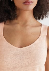 Persikofärgad linne med V-ringning, breda axelremmar och ett mjukt, något texturerat tyg. Enkel och minimalistisk estetik.