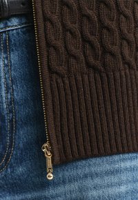 Close-up van een bruine kabelgebreide trui met een gouden rits over een zak van blauwe denim jeans met zichtbare riemlus en stiksels.