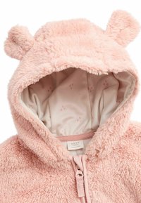 Haut de survêtement en polaire rose pour bébé avec des oreilles d'ours, texture douce, fermeture éclair à l'avant et doublure à motifs avec de petites empreintes de pattes.