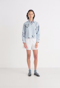 Kortärmad blårandig skjorta med svarta hjärttryck, i kombination med vita shorts i ögonmask. Modellen bär blå strumpor och svarta sandaler.