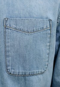 Hellblauer Denimstoff mit einer rechteckigen, abgesteppten Tasche mit einer spitzen Klappe, die eine glatte Textur und gleichmäßige Nähte zeigt.