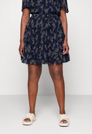 Marineblaues, geblümtes Kleid mit geschichteten Rüschen, Taillenschnürung und kurzen Ärmeln. Kombiniert mit hellbeigen Sandalen zum Hineinschlüpfen mit gerafften Details.