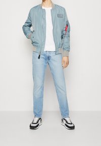 Light blue bomber jacket with grey cuffs, a patch on the chest, and a red zipper pocket. Worn over a white t-shirt and light blue jeans.