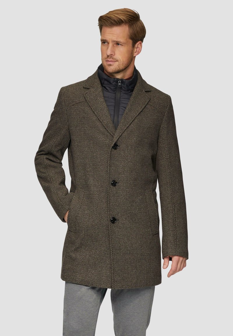 Hahnentritt-Wollmischung Mantel in Braun mit schwarzen Knöpfen, Reverskragen und seitlichen Taschen, über einer schwarzen, mit Reißverschluss versehenen Innentasche getragen.