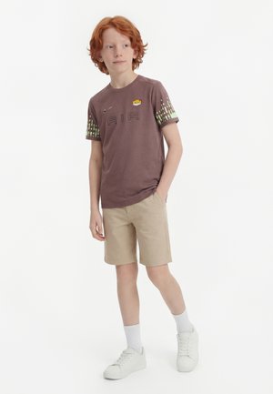 Garçon aux cheveux roux debout, une main dans la poche, portant un t-shirt graphique Nike Air marron, un short beige, des chaussettes blanches et des baskets blanches.