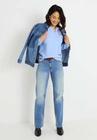 Suéter azul claro y esponjoso con un escote henley, combinado con jeans de tiro alto azul claro y una chaqueta vaquera colgada sobre los hombros.