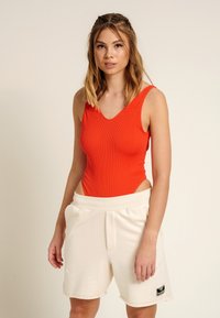 Roter, gerippter Bodysuit mit V-Ausschnitt und seitlichen Ausschnitten, kombiniert mit off-white Shorts mit elastischem Bund und Logo-Patch. Minimalistisches Design.