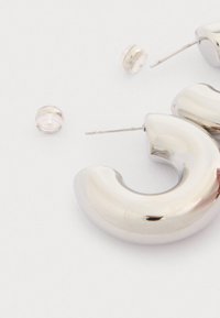 Cercei din argint în formă de cerc, cu o suprafață netedă și lucioasă. Include tortițe transparente și un design curbat, subliniind o estetică modernă.